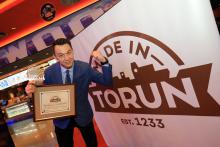 Zdjęcie zrobione podczas gali Made in Toruń. Laureat stoi na tle grafiki Made in Toruń. Prawą dłonią trzyma certyfikat, lewą zaciska i unosi do góry.