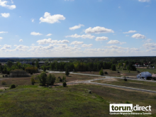 Widok ulicy Przelot i Świdnickiej w Toruniu z lotu ptaka. Asfaltowa droga pośród łąki. Na horyzoncie las i budynki