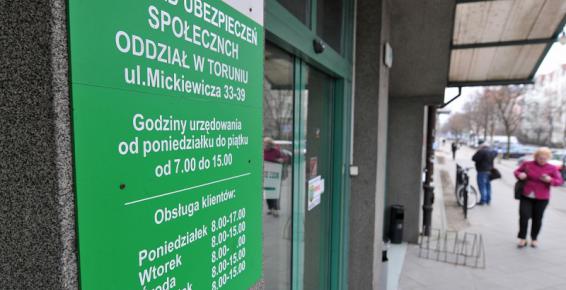 Fotografia przedstawiająca wejście do Zakładu Ubezpieczeń Społecznych przy ulicy Mickiewicza w Toruniu. Zielony szyld i drzwi wejściowe na pierwszym planie. W tle widoczny chodnik i przechodnie