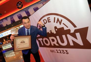 Laureat znaku pozuje z certyfikatem na tle banera z logiem Made in Toruń